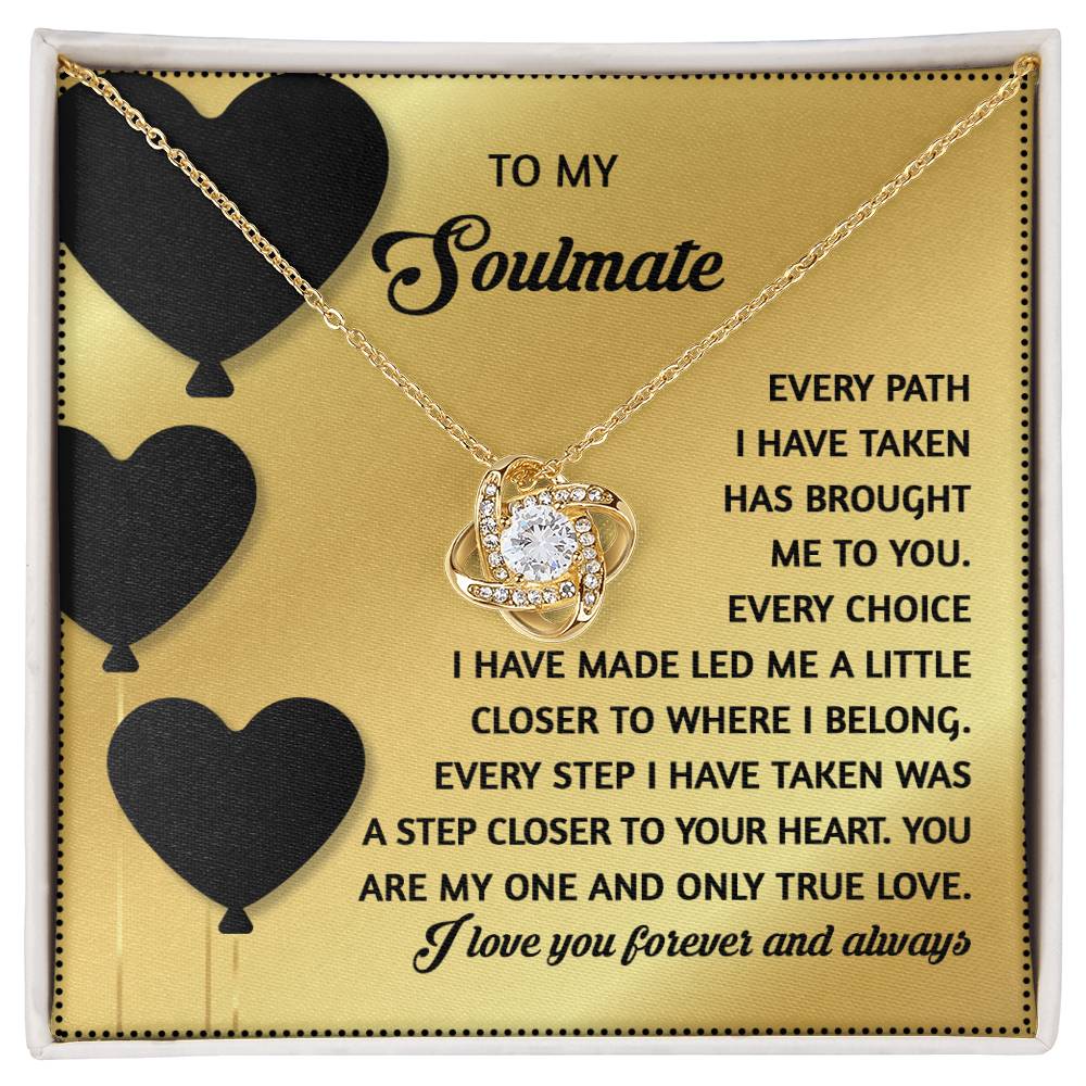 The "Soulmate-A Step Closer - Love Knot Necklace" features a gemstone heart pendant on a gift card with black hearts and text, expressing love and devotion. The elegant gold finish enhances its beauty.
