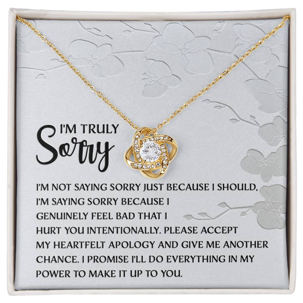 Introducing the Sorry-Feel Bad - Love Knot Necklace, featuring a pendant on a card with an apology message: "I'm truly sorry. I'm not saying sorry just because I should..." and continues with more heartfelt text. The necklace showcases a sparkling cubic zirconia crystal set in your choice of white gold or yellow gold finish, all against a light floral design background.