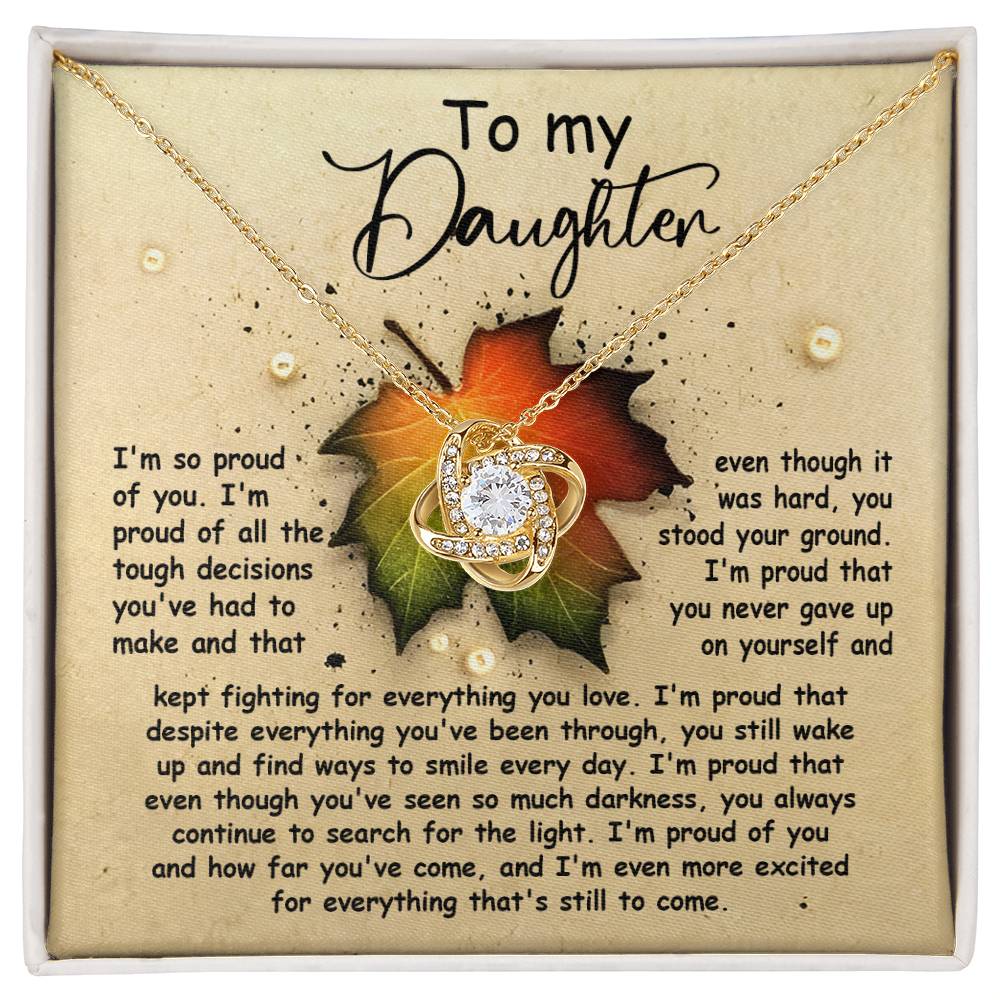 The "Daughter-Stood Your Ground - Love Knot Necklace" features a stunning gold finish and sparkling cubic zirconia crystals. It is elegantly displayed on a card bearing a heartfelt message of pride and admiration for your daughter's strength and perseverance, with a delicate leaf graphic subtly placed behind the pendant.