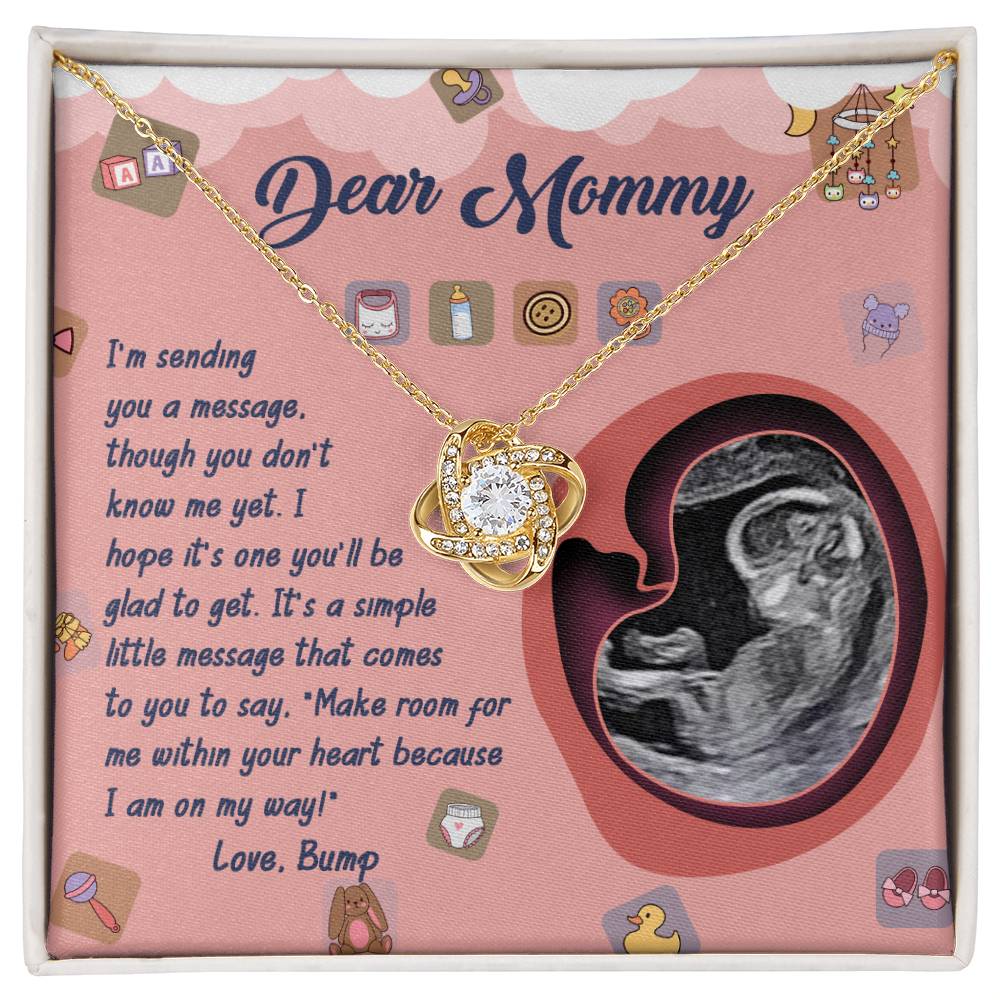 The "To Mom To Be, On My Way - Love Knot Necklace," adorned with cubic zirconia crystals, rests on a gold chain and comes with a heartfelt card featuring an ultrasound image and illustrations of baby items. This personalized gift beautifully captures the essence of love and anticipation.