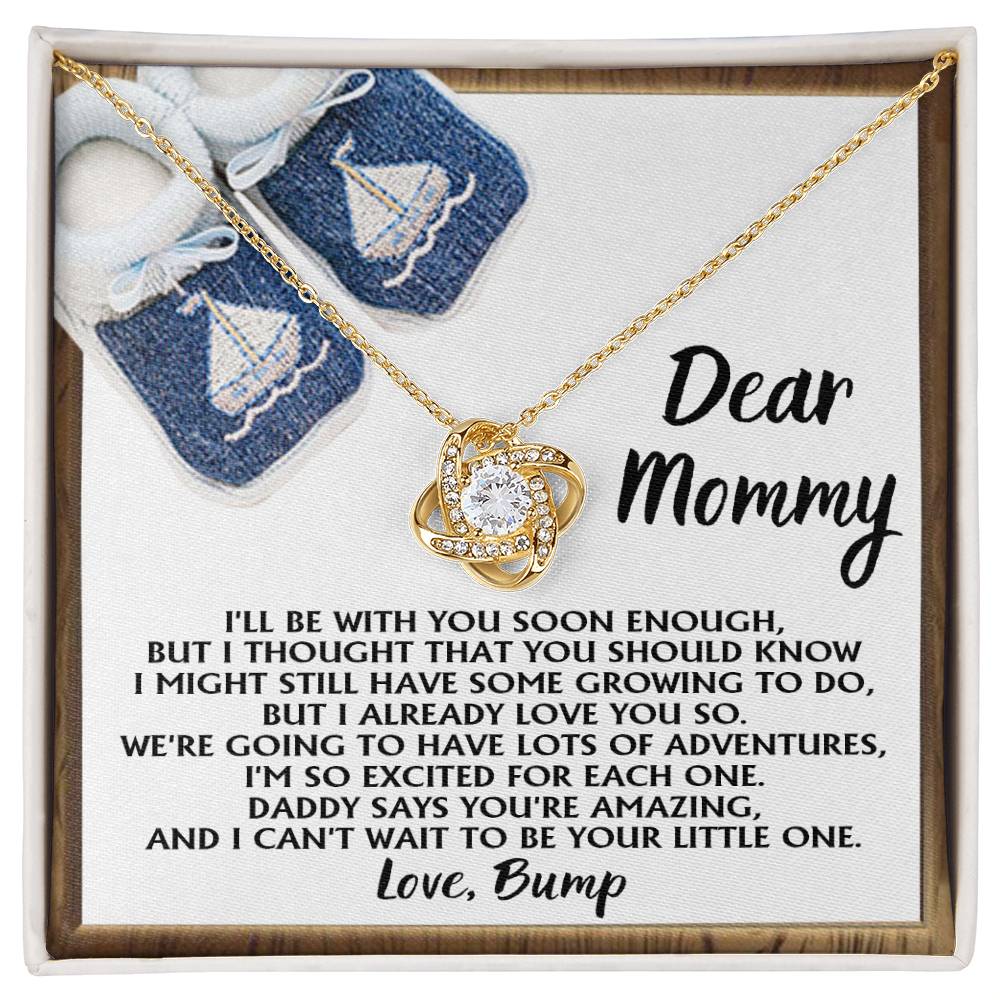 The "To Mom To Be, You're Amazing - Love Knot Necklace," featuring 14k white gold and cubic zirconia crystals, is presented on a card with a heartfelt message from an unborn child to the mother. In the background, there is a pair of blue baby booties adorned with white sailboats.