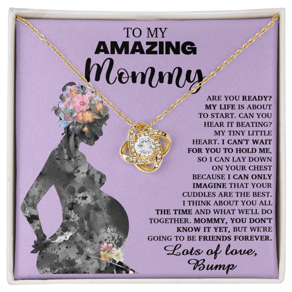 The "To Mom To Be, Tiny Little Heart - Love Knot Necklace," featuring a heart pendant, is laid over a printed message titled "To My Amazing Mommy," which showcases a silhouette of a pregnant woman adorned with floral patterns and premium cubic zirconia crystals.