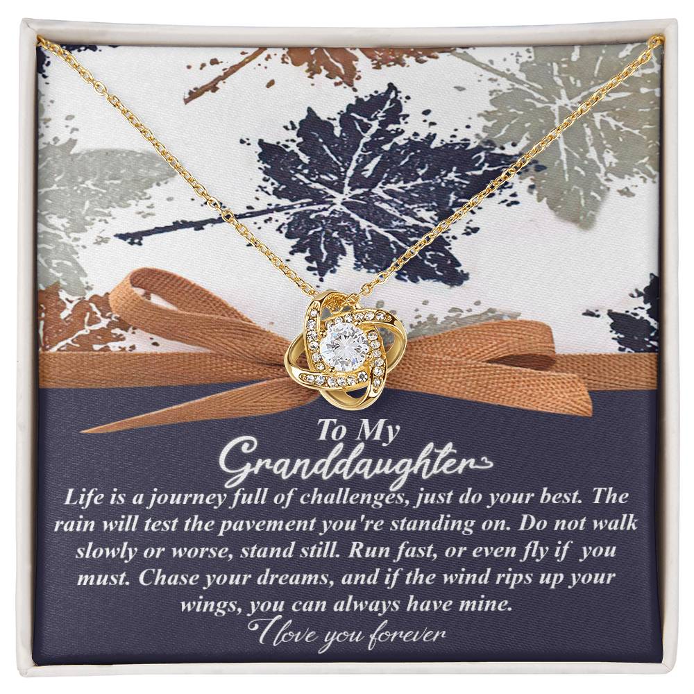 The Granddaughter-Have Mine - Love Knot Necklace, featuring a heart design, is beautifully placed over a card with a heartfelt message to a granddaughter and is elegantly presented in a decorative box. The necklace is crafted from white gold and adorned with sparkling cubic zirconia for added brilliance.