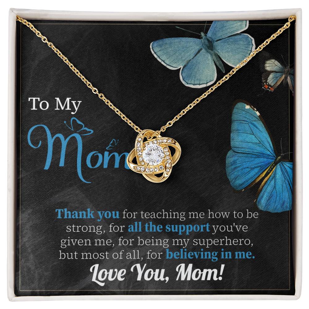 The "To Mom, Believing In Me - Love Knot Necklace" adorned with cubic zirconia crystals is displayed against a dark background featuring blue butterflies and a heartfelt note addressed to a mother, expressing gratitude and love.