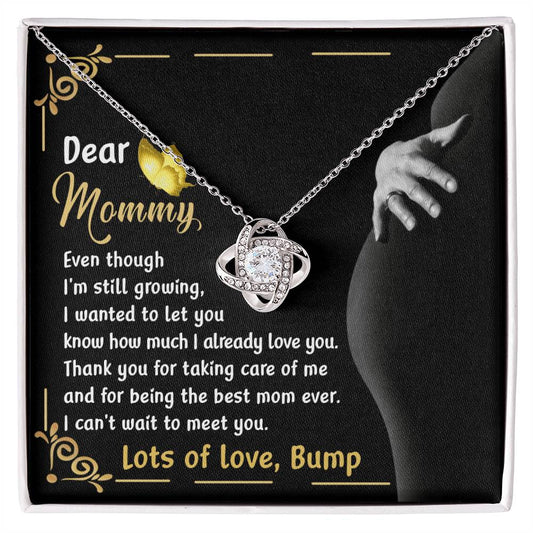 The "To Mama To Be, Best Mom Ever - Love Knot Necklace" comes in a gift box with a heartfelt message addressed to "Mommy." The note, signed "Lots of love, Bump," expresses love and gratitude from an unborn child. The necklace features an elegant intertwined design adorned with cubic zirconia crystals and is available in either a white gold or yellow gold finish.