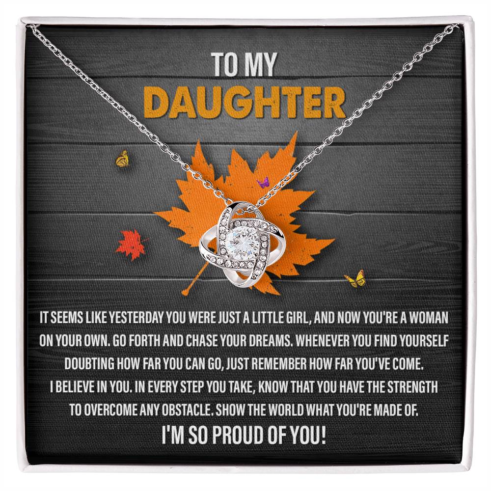 The Daughter-You Can Go - Love Knot Necklace, crafted in white gold and embellished with cubic zirconia, is showcased against a box adorned with an orange maple leaf and a touching message to a daughter about pursuing dreams and overcoming obstacles. This text conveys pride and heartfelt encouragement.