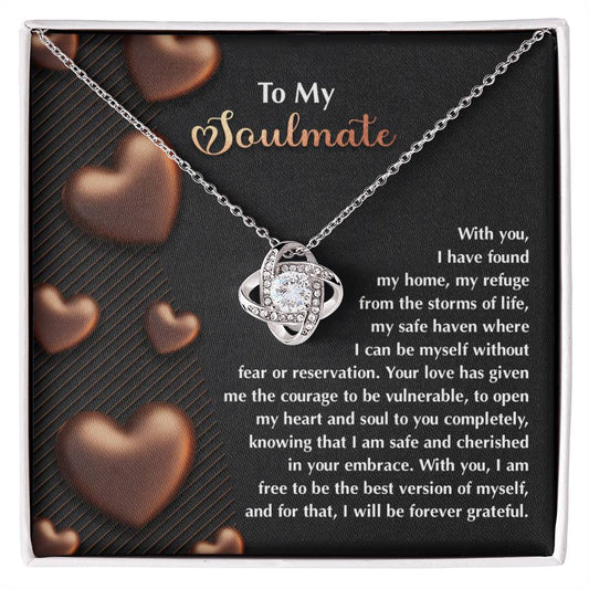 The Soulmate-Best Version Love Knot Necklace features a silver intertwined heart with a cubic zirconia pendant in a box reading "To My Soulmate." The gold finish highlights the heartfelt love and gratitude message on a black and brown heart-adorned background.
