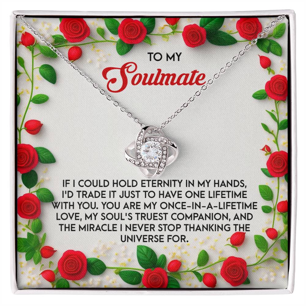 The Soulmate-One Lifetime - Love Knot Necklace, in white or yellow gold finish, is elegantly presented in a jewelry box with cubic zirconia crystals. It includes a heartfelt message about love and soulmates, bordered by red roses and lush green leaves.