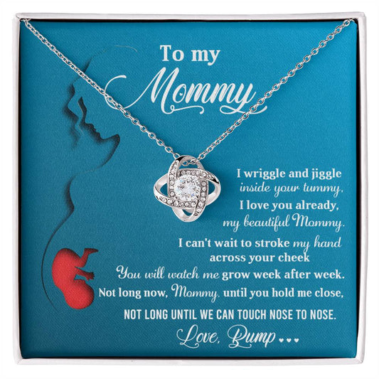 The "To Mom To Be, Nose To Nose - Love Knot Necklace" comes on a blue card with a heartfelt message from an unborn baby to their mother, expressing love and excitement to meet. Crafted in 14k white gold and adorned with sparkling cubic zirconia crystals, the necklace is displayed beside a card featuring the silhouette of a pregnant woman with her baby inside.