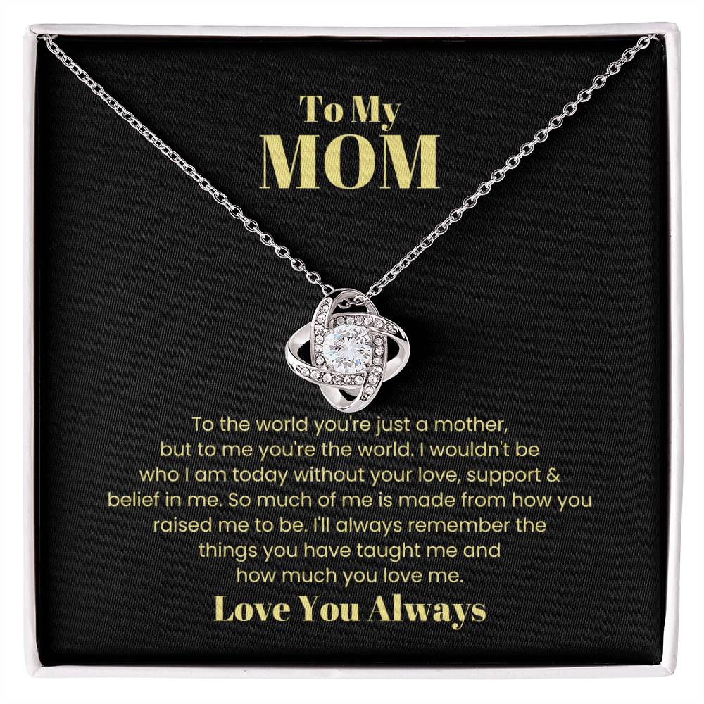 The "To My Mom, To The World You Are Just A Mother - Love Knot Necklace" features an intertwined pendant embellished with cubic zirconia crystals, set against a black background alongside a heartfelt message to a mother that conveys love, gratitude, and the significance of her support.