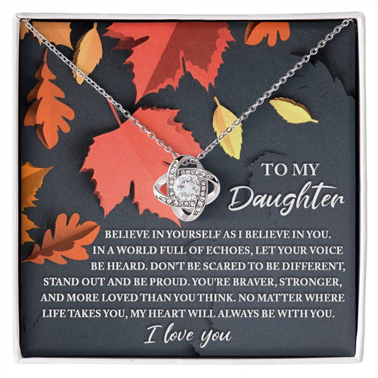 A Daughter-To Be Different - Love Knot Necklace, elegantly adorned with cubic zirconia crystals, rests on a black and orange leaf-patterned background. It is accompanied by a heartfelt message to a daughter about bravery, strength, and love—making it the perfect personalized gift.