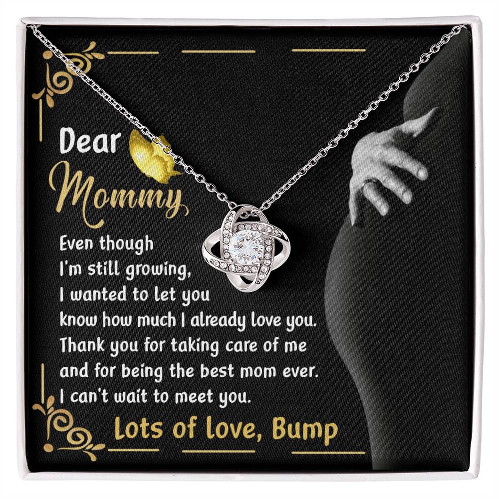 The "To Mama To Be, Best Mom Ever - Love Knot Necklace" features a heart pendant adorned with cubic zirconia crystals and comes with a touching card that conveys a heartfelt message from an unborn baby to its mother, expressing love and gratitude. The background image of a pregnant belly enhances this sentimental moment.