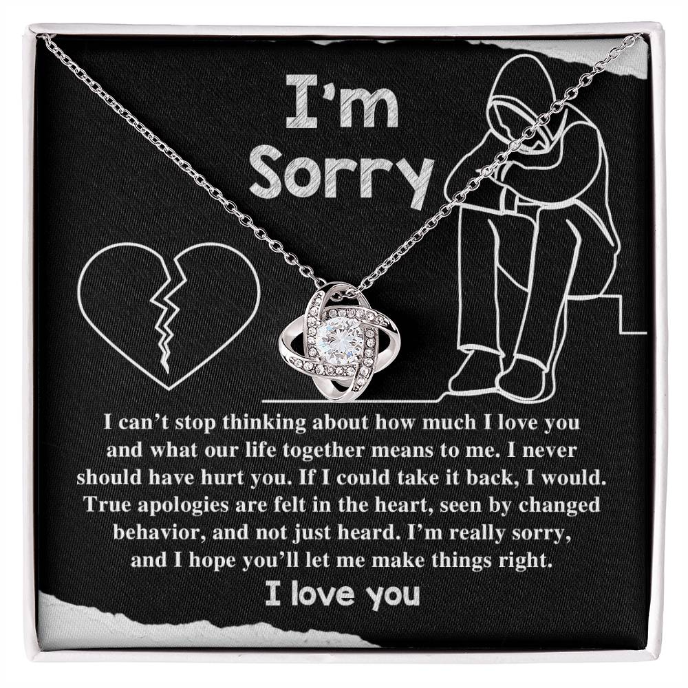 The "Sorry-Not Just Heard - Love Knot Necklace" features an apology message on a black background. The heartfelt text expresses regret and a promise to change, accompanied by illustrations of a heart and a person sitting with their head down. Cubic zirconia crystals add a touch of elegance, with "I love you" written at the bottom.