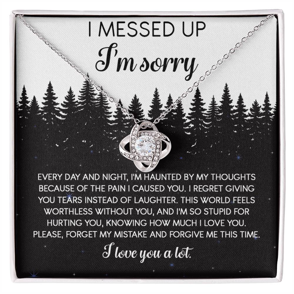 The Sorry-By My Thoughts - Love Knot Necklace, featuring a heart-shaped pendant adorned with cubic zirconia crystals, is displayed on a card with an apology message. The card reads: "I messed up. I'm sorry. Every day and night, I'm haunted...I love you a lot," set against a serene forest background.
