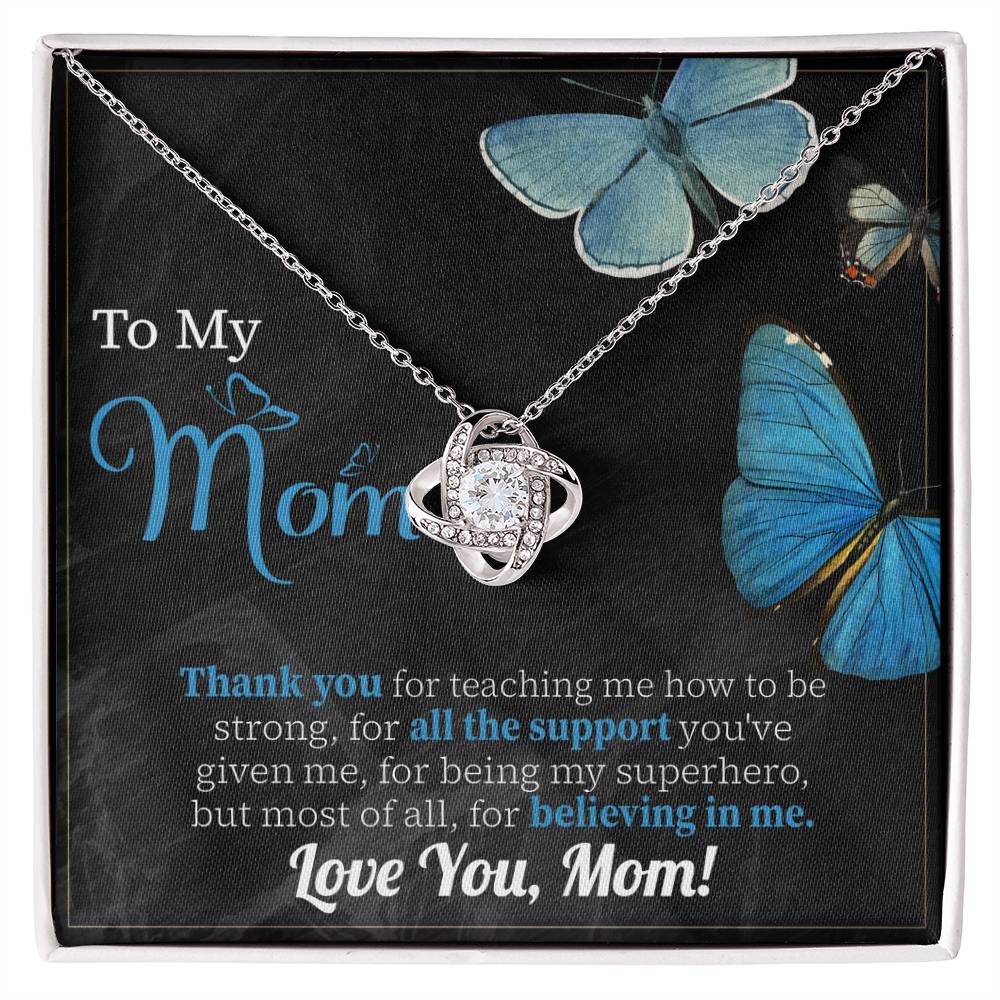 The "To Mom, Believing In Me - Love Knot Necklace" features an elegant intertwined design and comes on a beautifully decorated card with blue butterflies and a touching message for "Mom." The note expresses gratitude for her support, strength, and love, concluding with "Love You, Mom!" This personalized gift sparkles with cubic zirconia crystals.