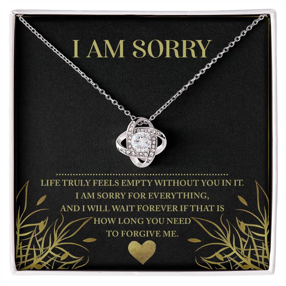 A Sorry-Without You - Love Knot Necklace in silver, featuring an intricate interwoven knot pendant adorned with sparkling cubic zirconia, is showcased against a black background. The packaging bears a heartfelt message in white and gold lettering: "I AM SORRY" at the top, followed by, "Life truly feels empty without you in it. I am sorry for everything, and I will wait forever if that is how long you need.