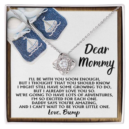 The "To Mom To Be, You're Amazing - Love Knot Necklace," graced with sparkling cubic zirconia crystals, is showcased in front of a greeting card addressed to "Mommy" and signed "Love, Bump," filled with a heartfelt message. In the background, baby shoes featuring sailboat designs add a whimsical touch.