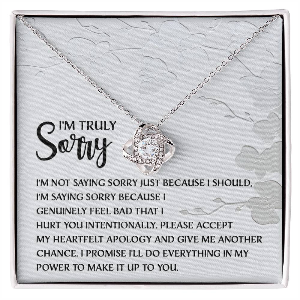 Introducing the Sorry-Feel Bad - Love Knot Necklace, featuring a pendant on a card with an apology message: "I'm truly sorry. I'm not saying sorry just because I should..." and continues with more heartfelt text. The necklace showcases a sparkling cubic zirconia crystal set in your choice of white gold or yellow gold finish, all against a light floral design background.