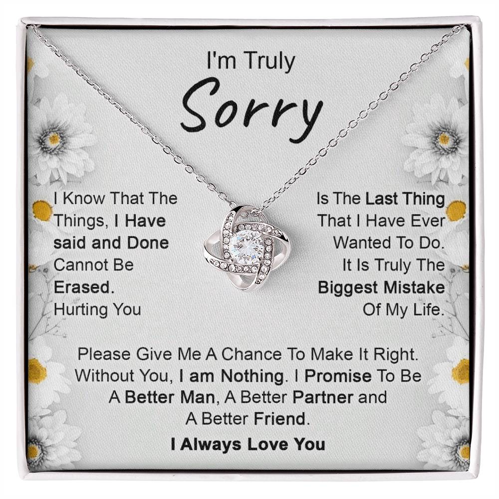 The "I’m Truly Sorry - Love Knot Necklace," featuring a pendant adorned with cubic zirconia crystals, rests on a heartfelt message that reads, "I'm Truly Sorry." The text conveys remorse for past actions and a commitment to becoming a better person, partner, and friend. The background is decorated with delicate floral designs.