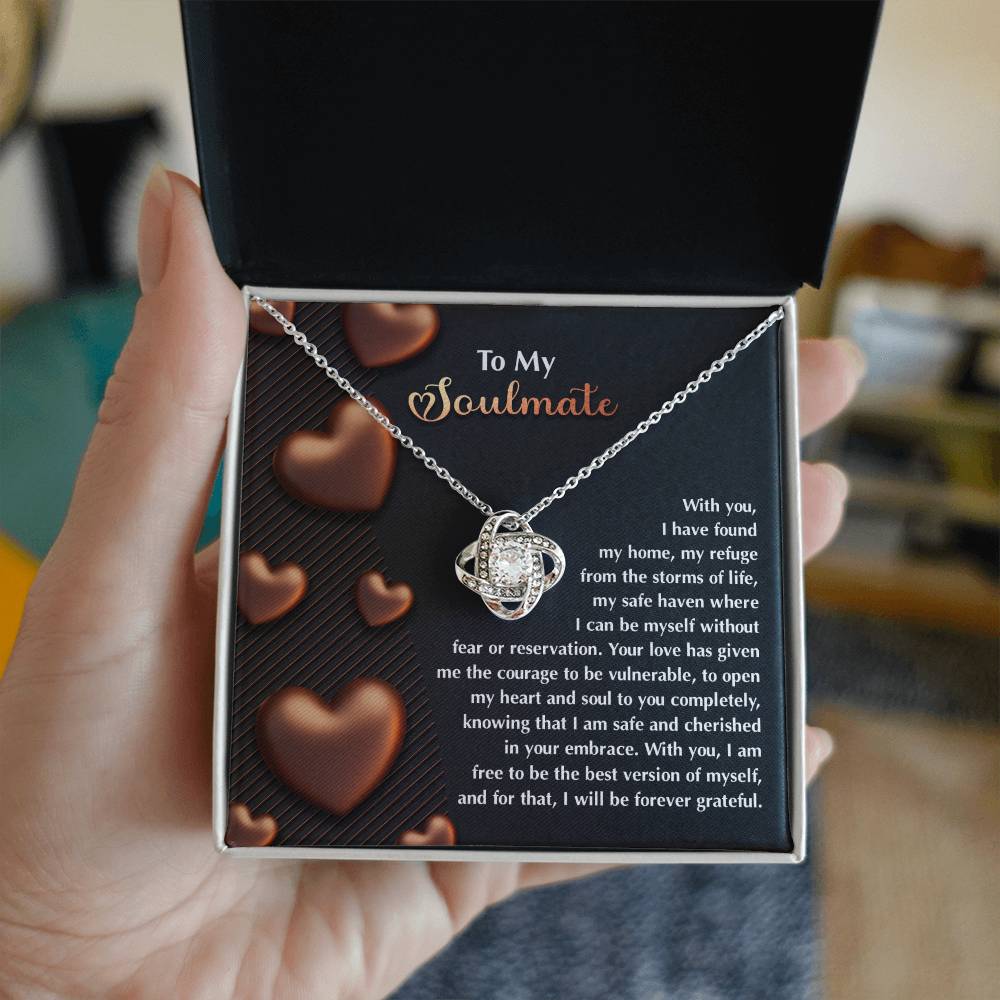A hand opens a box to reveal the Soulmate-Best Version - Love Knot Necklace, crafted in 14k white gold and adorned with Cubic Zirconia. The inside of the lid displays a heartfelt message with heart images on a deep brown background.