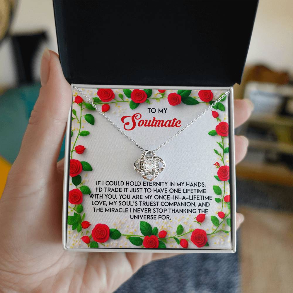 A hand unveils the Soulmate-One Lifetime - Love Knot Necklace, embellished with cubic zirconia crystals. The romantic message is enhanced by a floral design featuring red roses, complementing the necklace's elegant gold finish.
