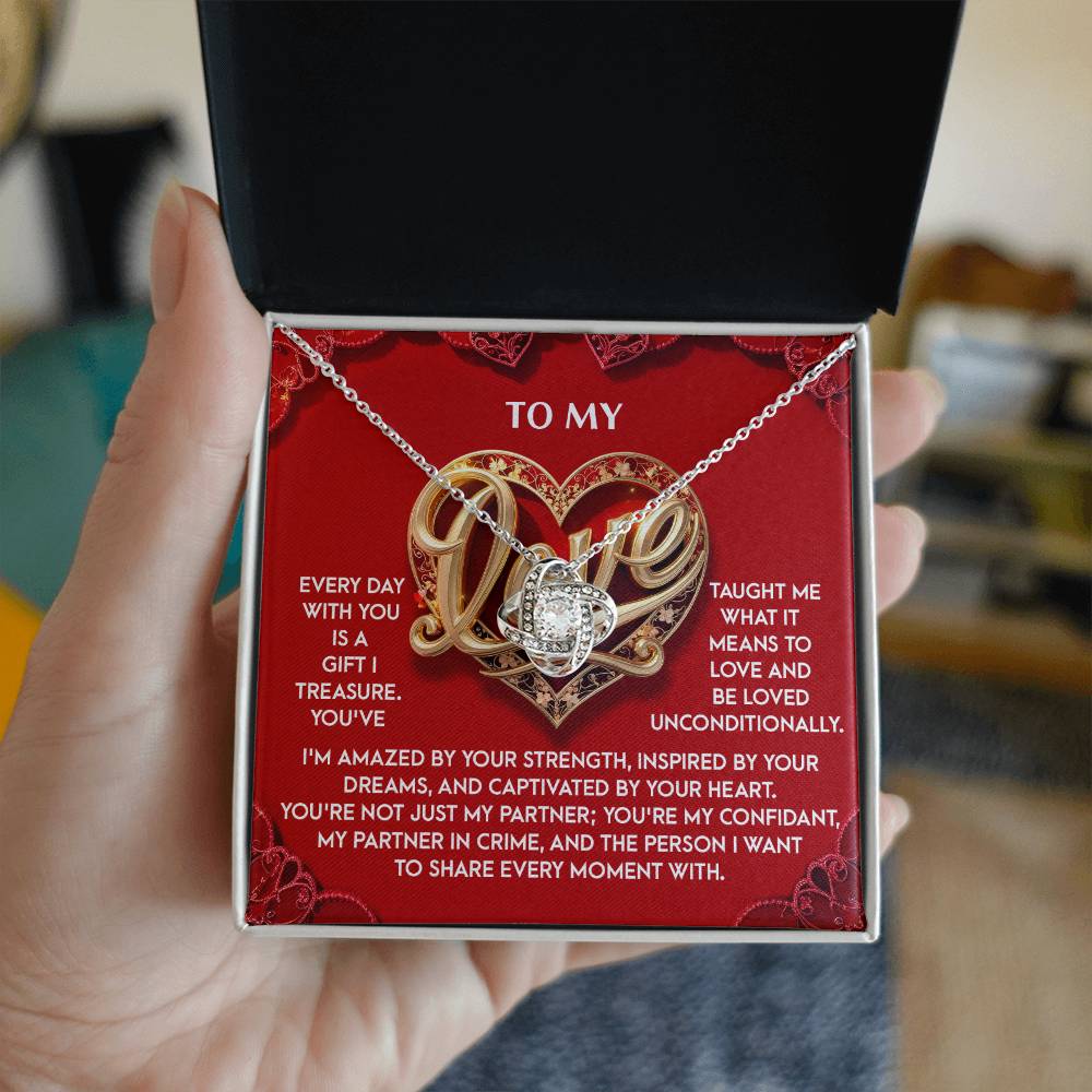 A person holds an open jewelry box showing the My Love-Every Moment - Love Knot Necklace, adorned with shimmering cubic zirconia crystals, against a background displaying a romantic message.