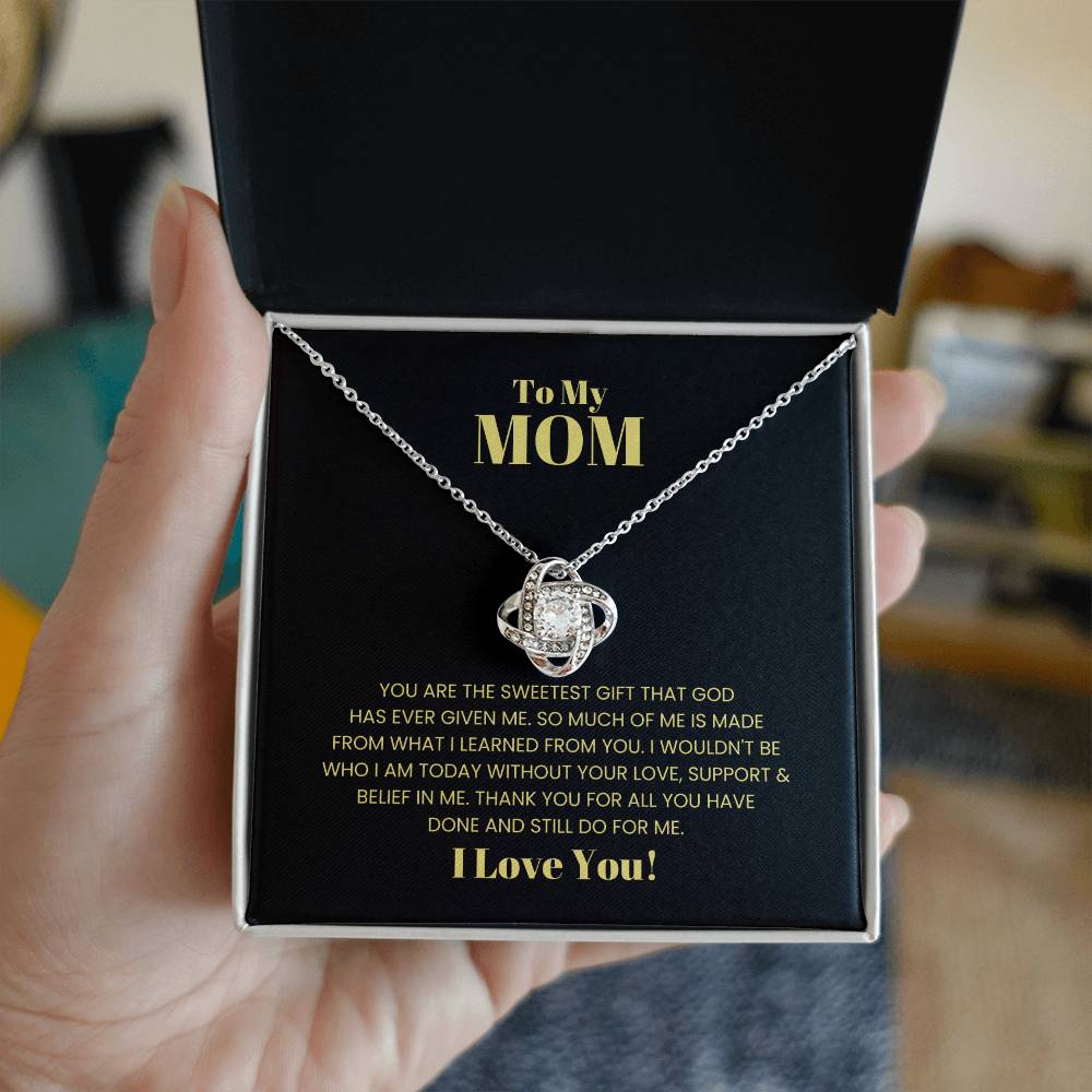 A hand holds an open jewelry box containing the "To My Mom, You Are The Sweetest Gift - Love Knot Necklace," adorned with premium cubic zirconia crystals. The box's lid bears an emotional message dedicated to "Mom," expressing deep gratitude and love, making it the perfect personalized gift.