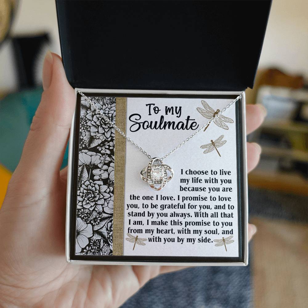 A hand holds an open jewelry box containing the exquisite "Soulmate-Make This Promise - Love Knot Necklace" along with a message: "To my soulmate," followed by a heartfelt promise of love, gratitude, and commitment, all adorned with floral patterns and sparkling cubic zirconia crystals.