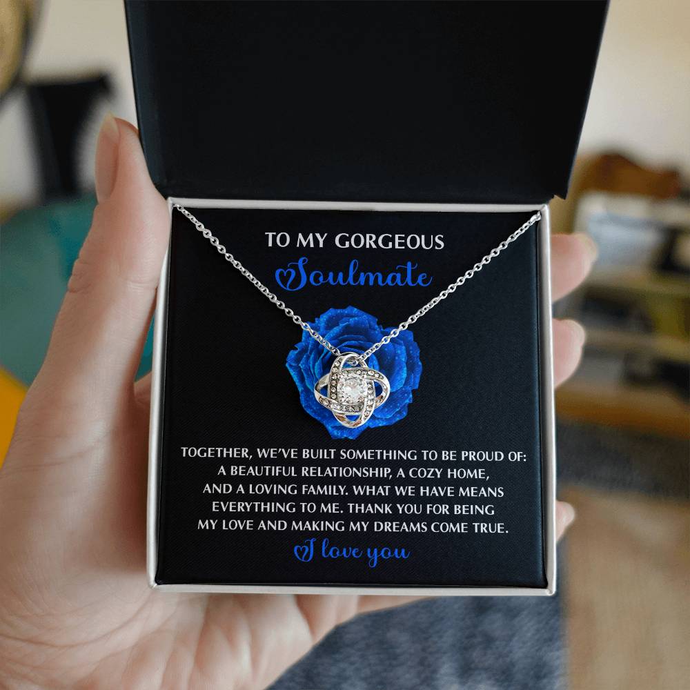 A hand holds a box with a shimmering Soulmate-Dreams Come True - Love Knot Necklace in white gold, adorned with cubic zirconia. The message states: "To my gorgeous soulmate, Together, we've built something to be proud of," and is accentuated by a decorative blue rose.