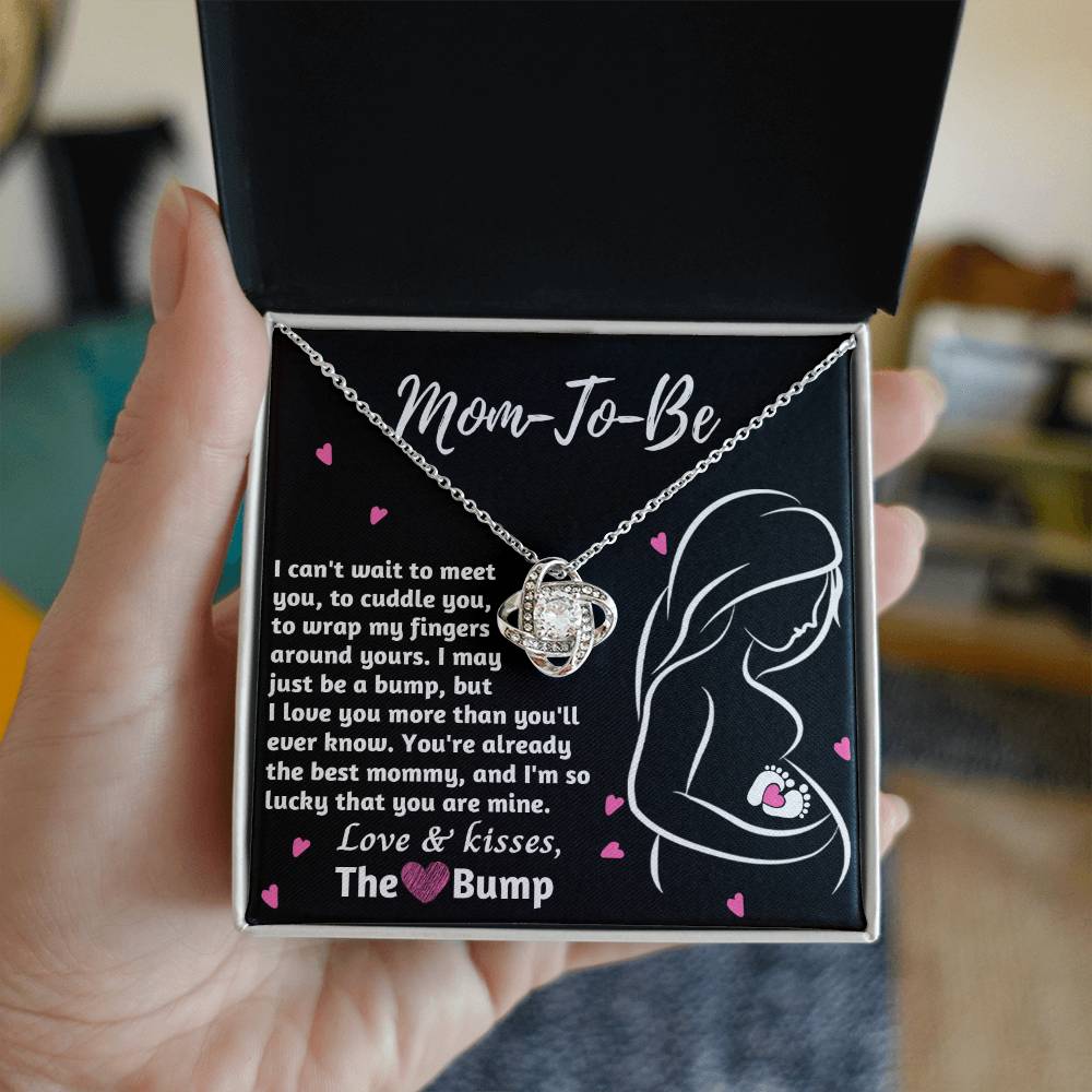 A hand holds a gift box containing a personalized To Mama To Be, The Best Mommy - Love Knot Necklace with a pendant and a message for an expectant mother from "the bump" by ShineOn Fulfillment.