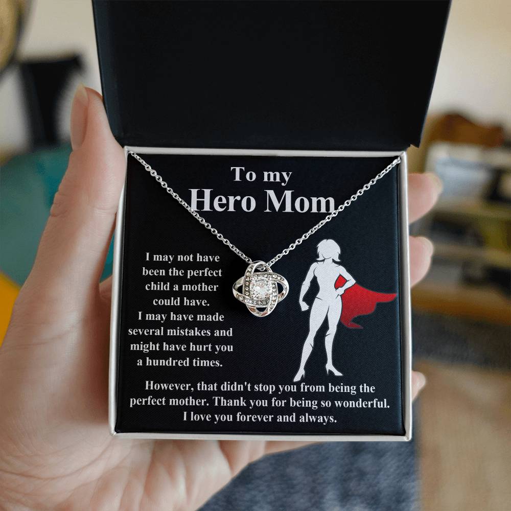 A hand holds an open jewelry box containing a stunning "To Mom, The Perfect Mother - Love Knot Necklace" adorned with cubic zirconia crystals and a note that reads, "To my Hero Mom" accompanied by a heartfelt message expressing gratitude for her enduring love and support.