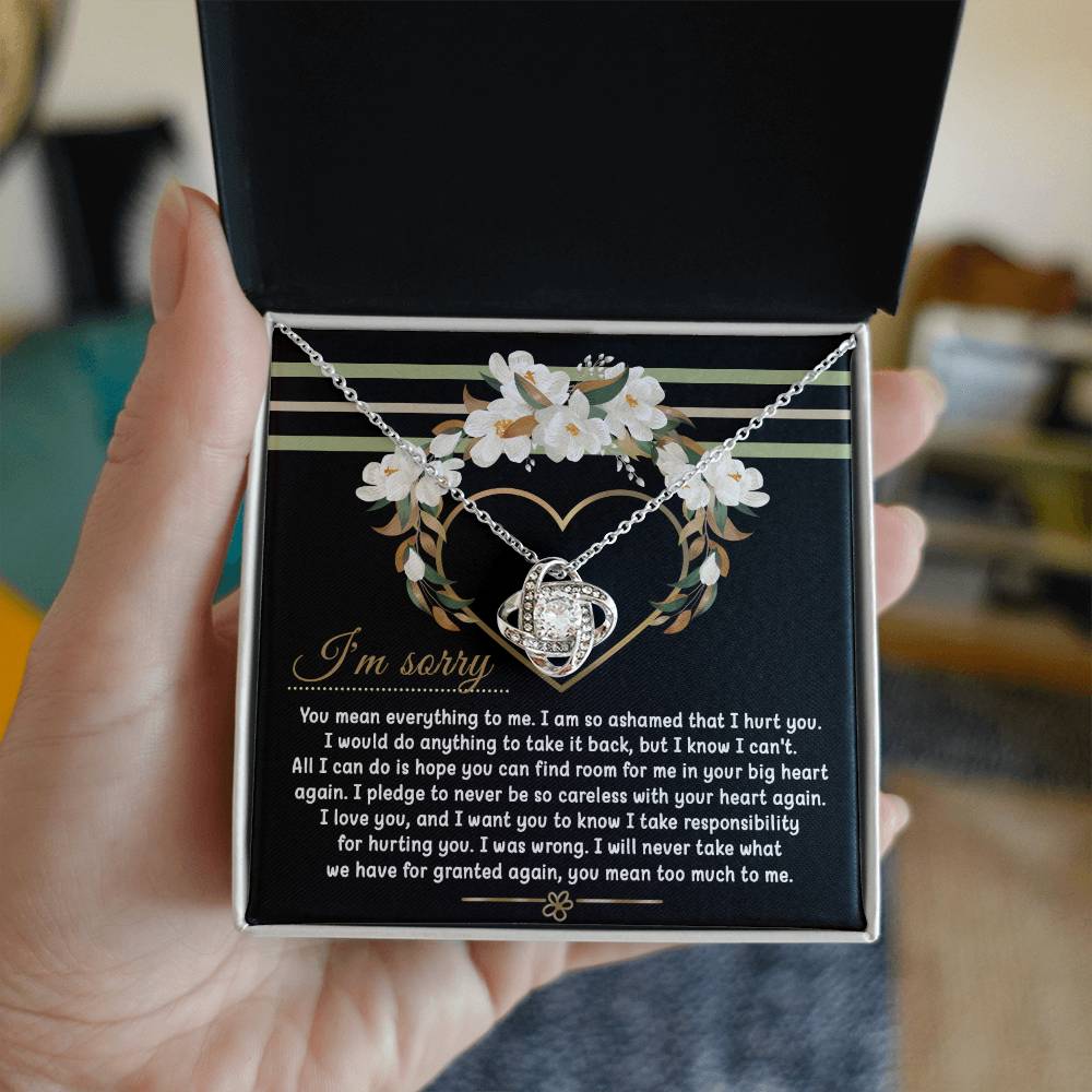 Introducing the Sorry-Big Heart - Love Knot Necklace: a heart-shaped pendant nestled in an elegant box, complete with an apology message expressing love and regret for causing hurt. The backdrop showcases a beautiful floral heart design, while the pendant sparkles with cubic zirconia crystals. Personalize your necklace by choosing between a white gold or yellow gold finish.