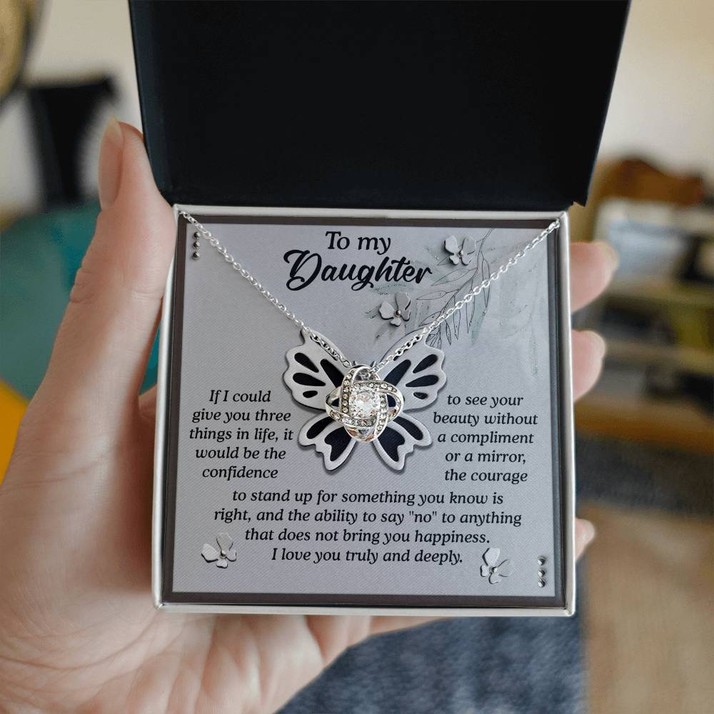 A person holds an open box containing the Daughter-See Your Beauty - Love Knot Necklace, a butterfly-shaped piece adorned with cubic zirconia crystals, accompanied by a message that reads, "To my Daughter," with additional text beneath it.