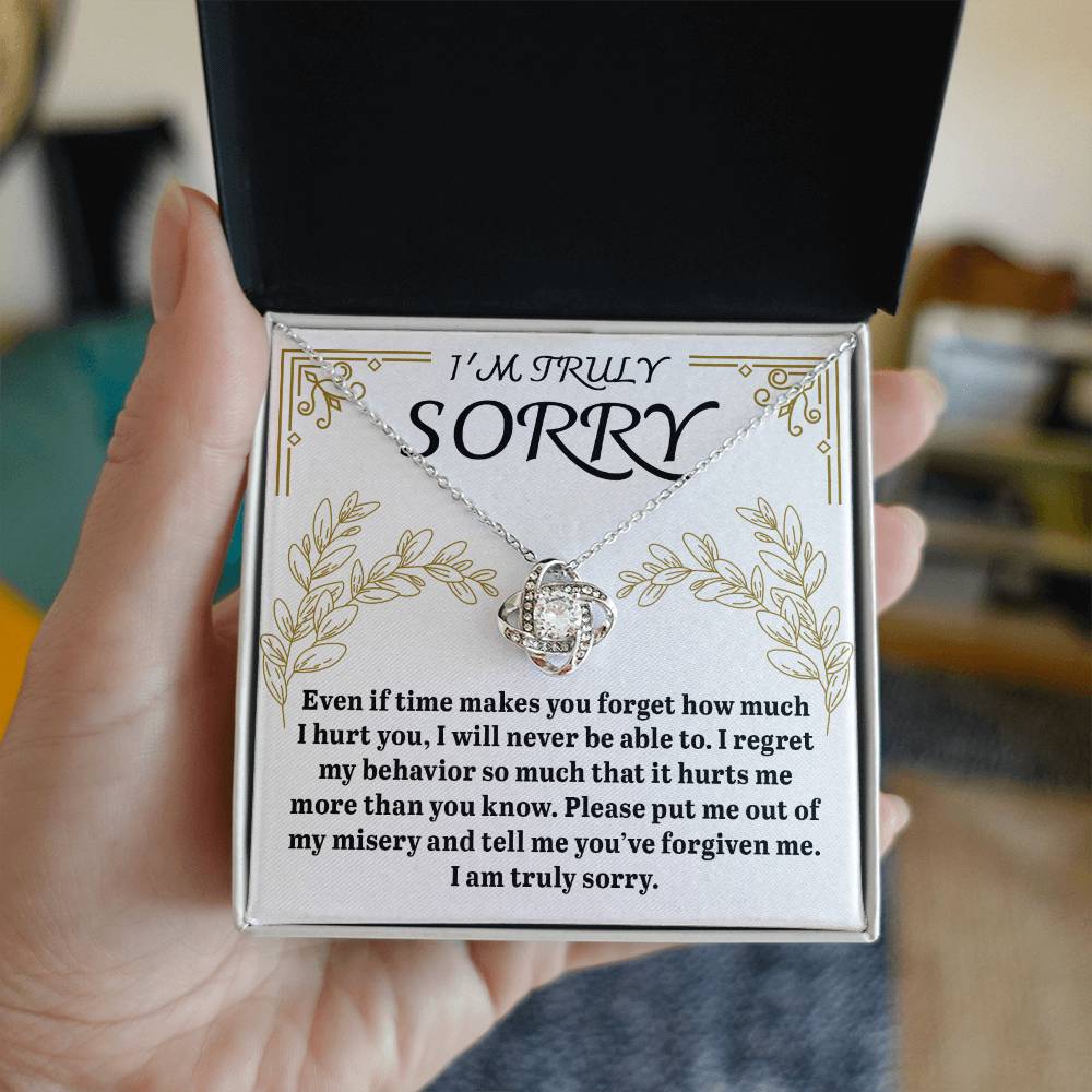 A hand holds an open box containing a Sorry-It Hurts Me - Love Knot Necklace adorned with cubic zirconia crystals, along with a card that expresses a heartfelt apology and a sincere request for forgiveness.