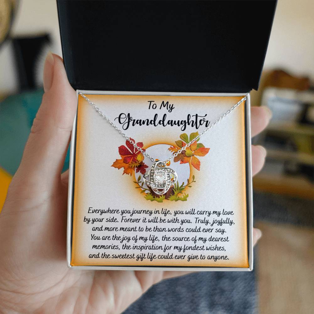An open gift box, held in hand and surrounded by vibrant autumn leaves, showcases the Granddaughter-By Your Side - Love Knot Necklace featuring a gold finish and shimmering cubic zirconia, alongside a heartfelt message for a granddaughter.