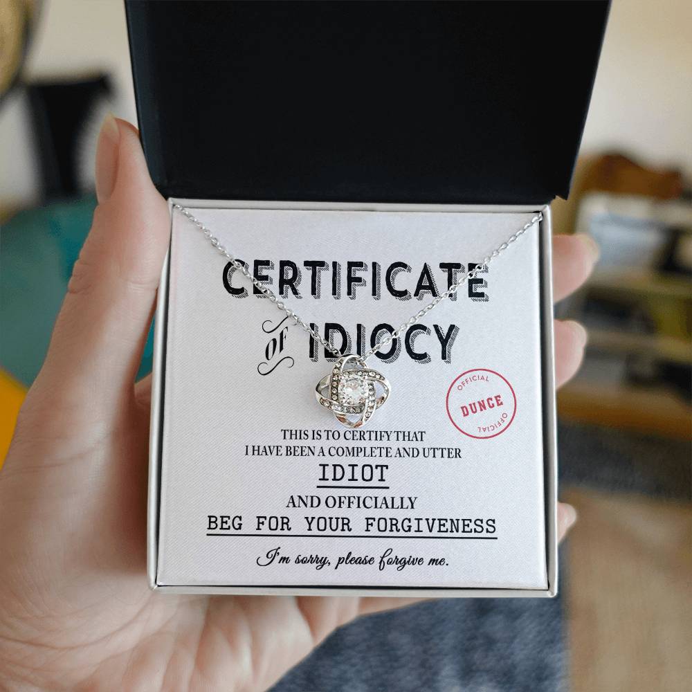 A hand holding an open jewelry box reveals a "Sorry-Certificate Of Idiocy - Love Knot Necklace" with a gold finish, adorned with cubic zirconia crystals. Inside lies a certificate labeled "Certificate of Idiocy," apologizing for being an idiot and begging for forgiveness.