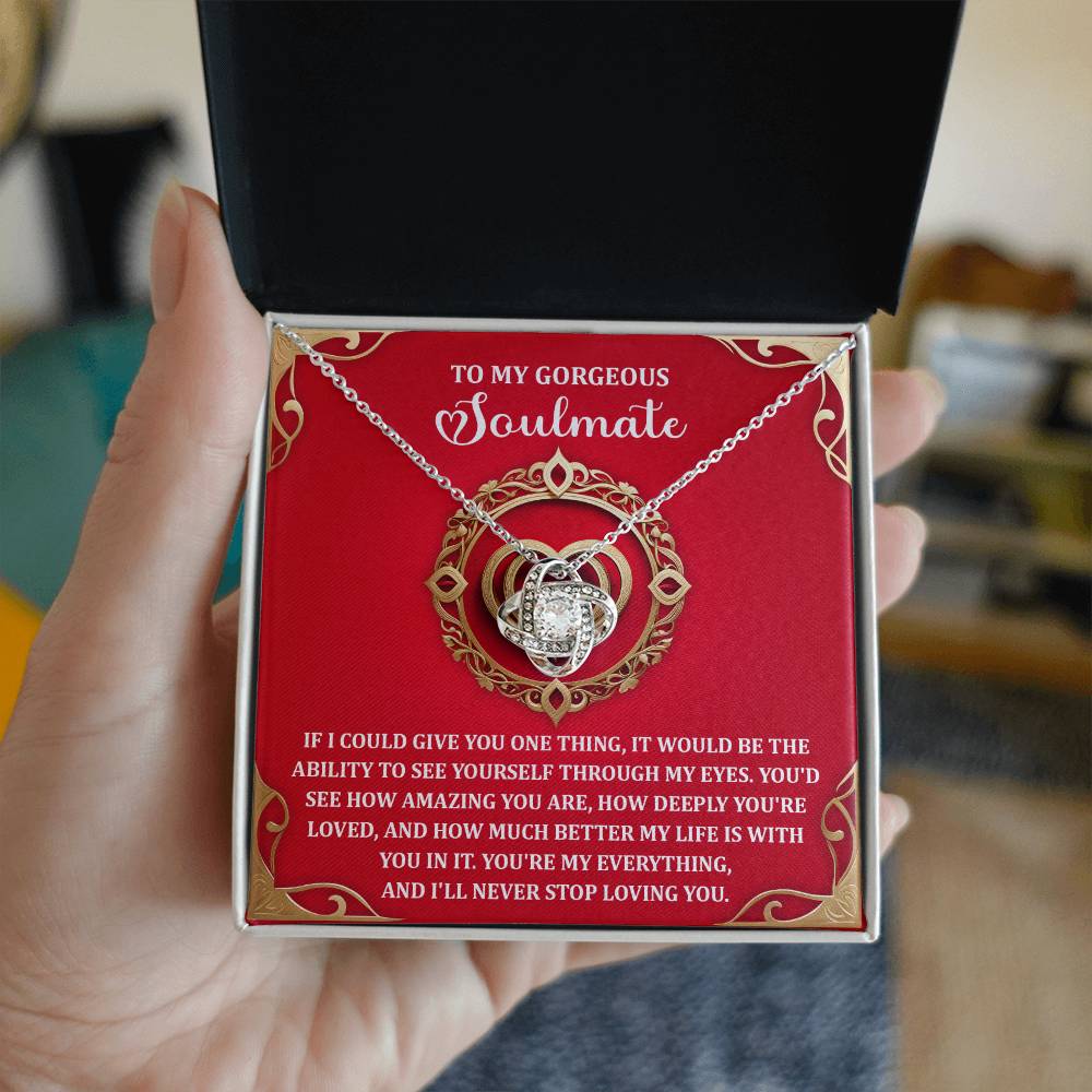 A hand holds an open jewelry box displaying the Soulmate-Through My Eyes - Love Knot Necklace adorned with sparkling cubic zirconia crystals. The box reads a romantic message to a "soulmate" on a rich red background, making it the perfect personalized gift.