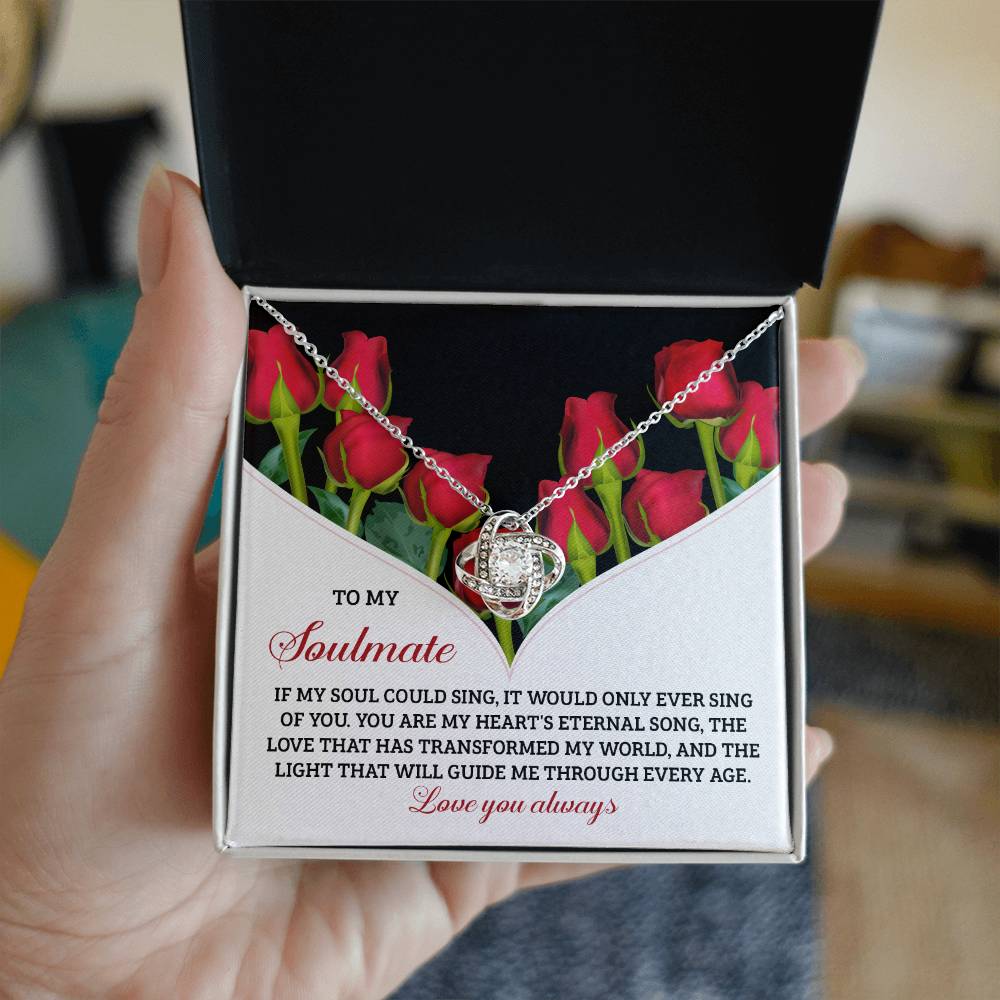 A hand opens a gift box containing the Soulmate-Eternal Song - Love Knot Necklace, featuring shimmering cubic zirconia. The box is decorated with red roses and heartfelt text of love and devotion to a soulmate.