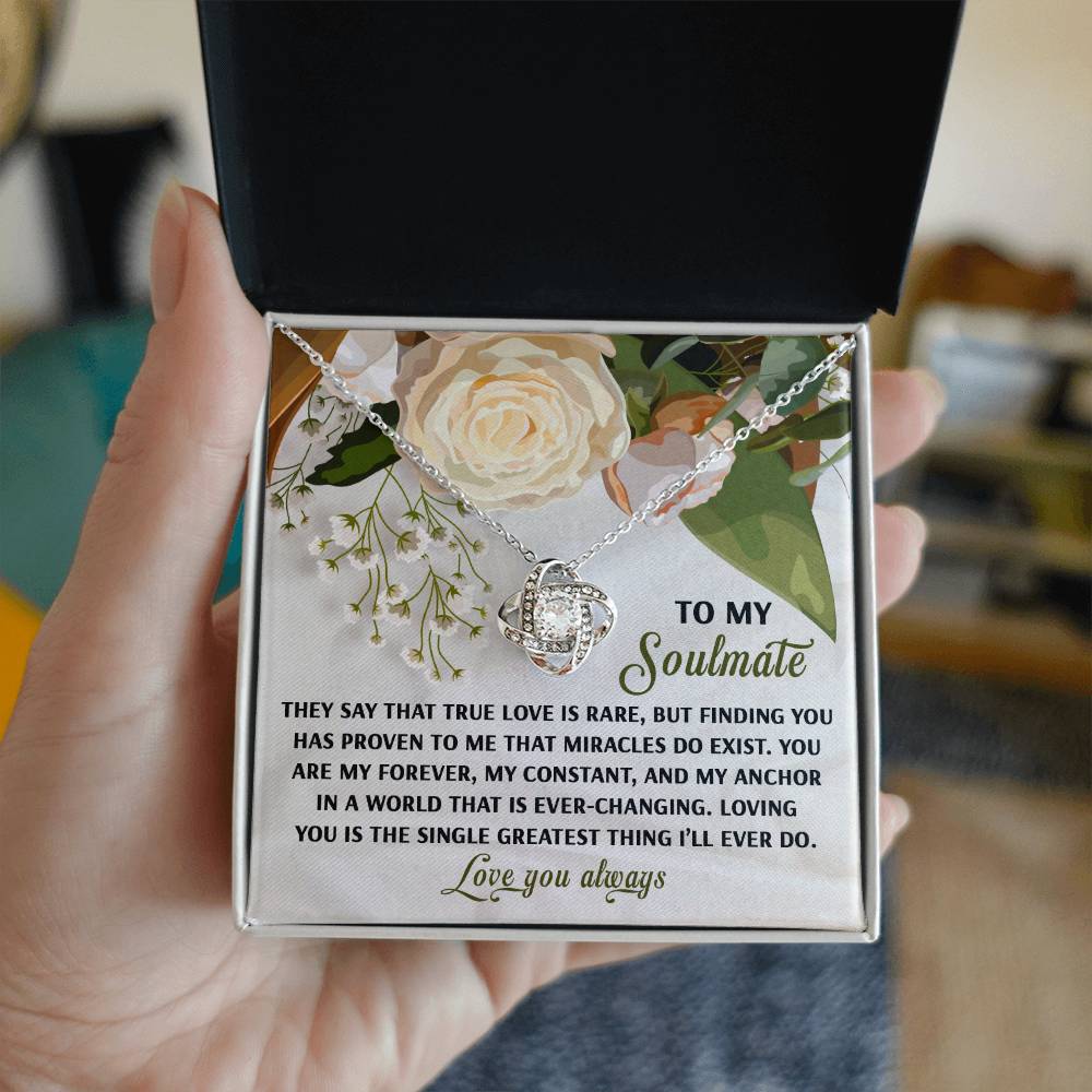 A hand holds an open jewelry box with a floral lid and heartfelt message, revealing the Soulmate-Greatest Thing Love Knot Necklace. The necklace, featuring cubic zirconia crystals, is available in white or yellow gold finishes.