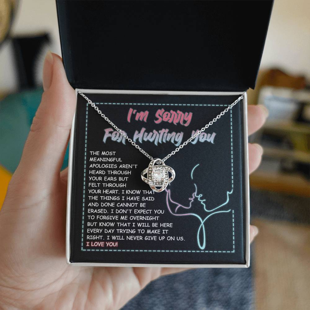 A 14k white gold Sorry-Never Give Up - Love Knot Necklace with an interlocked pendant set with cubic zirconia crystals sits on a note that reads, "I'm sorry for hurting you," accompanied by an apology message expressing regret, commitment to make things right, and affirming love.