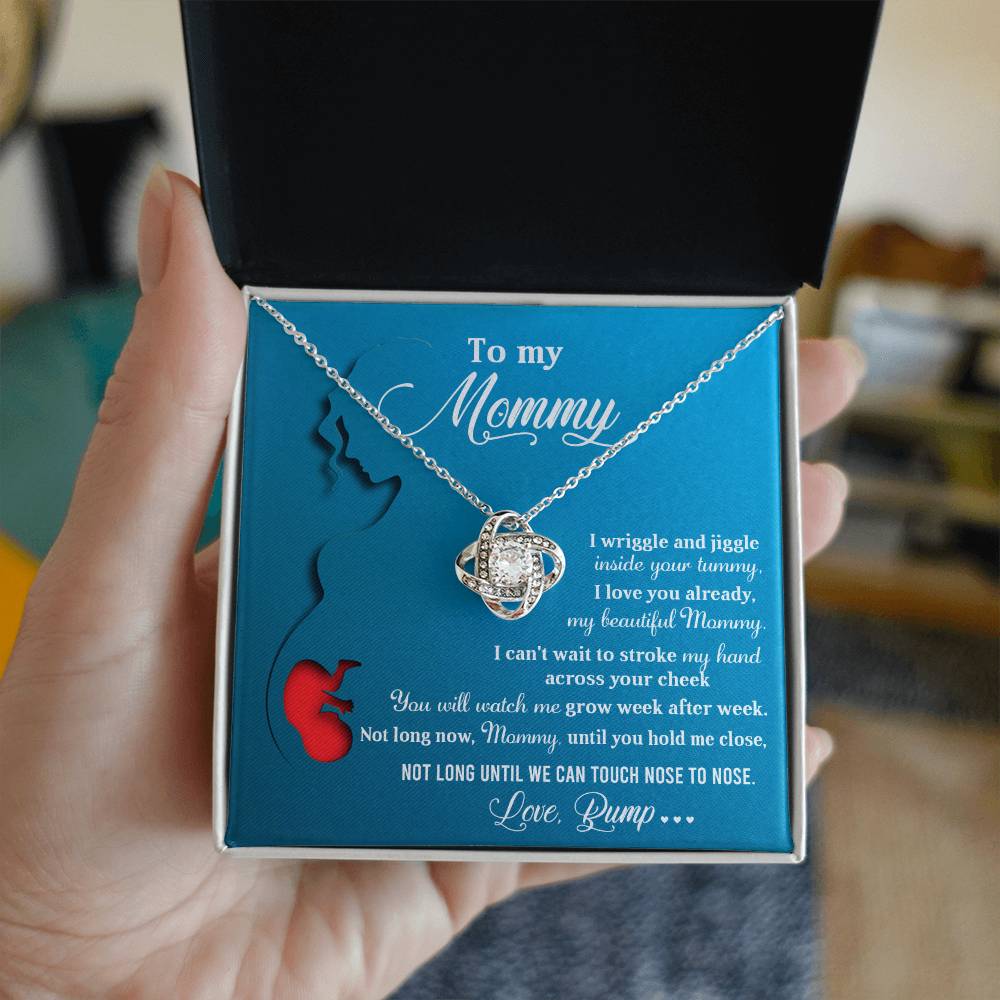 A hand holds an open box showcasing the "To Mom To Be, Nose To Nose - Love Knot Necklace," a stunning 14k white gold piece adorned with cubic zirconia crystals, in front of a heartfelt message to a mother from an unborn baby on a blue background card.