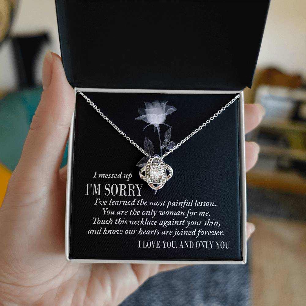 A silver Sorry-Painful Lesson - Love Knot Necklace on a dark background, adorned with shimmering cubic zirconia crystals. The text includes an apology, a statement of love, and an affirmation of unity.