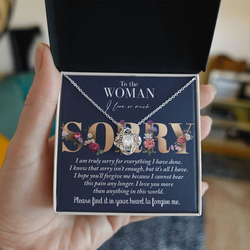 A person holds an open jewelry box containing a stunning "Sorry-Bear This Pain - Love Knot Necklace" adorned with cubic zirconia crystals. The box lid reads, "To the woman I love so much," followed by a detailed apology note.
