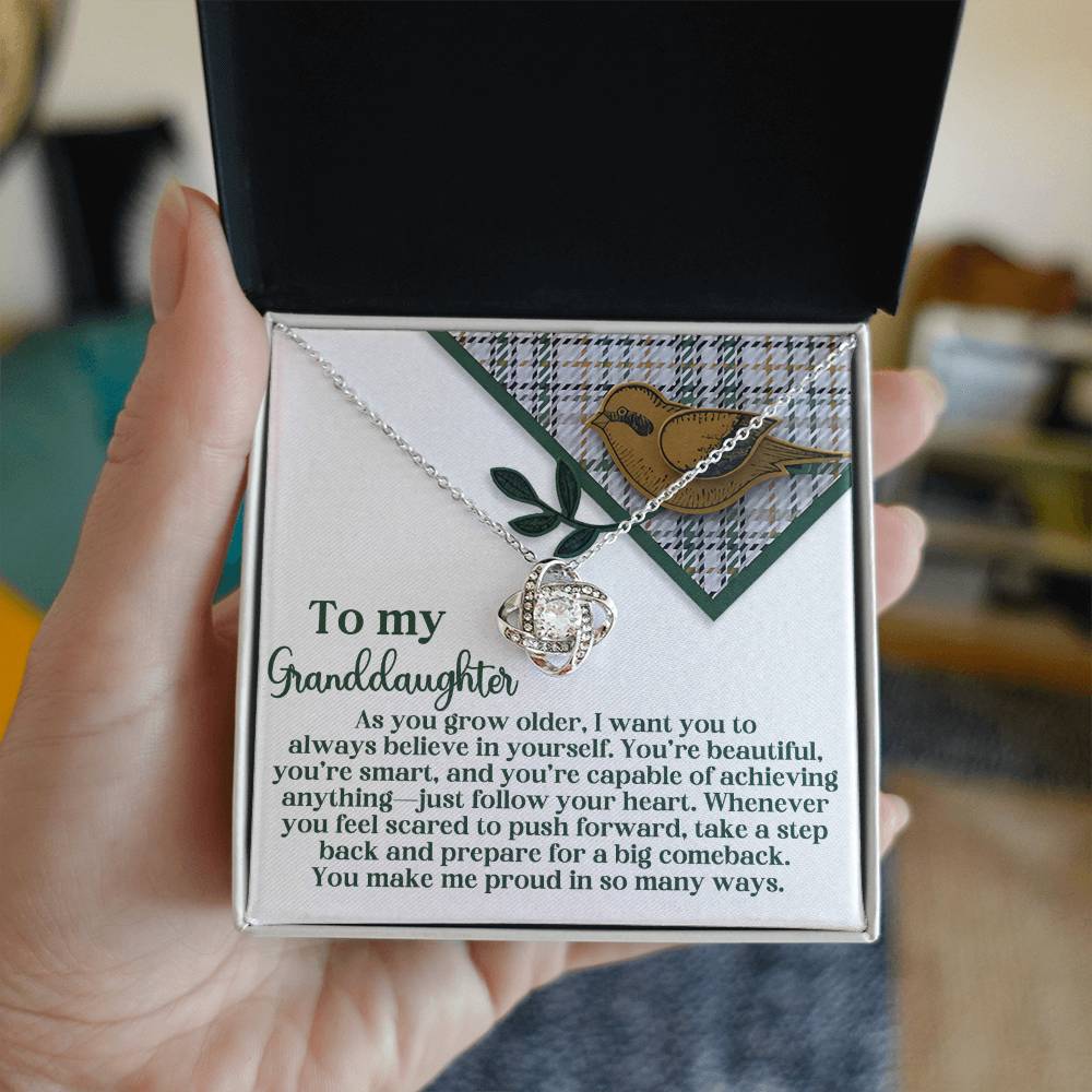 A person holds an open jewelry box displaying the exquisite Granddaughter-A Big Comeback - Love Knot Necklace, featuring cubic zirconia and a gold finish, accompanied by a card with an inspirational message for their granddaughter.