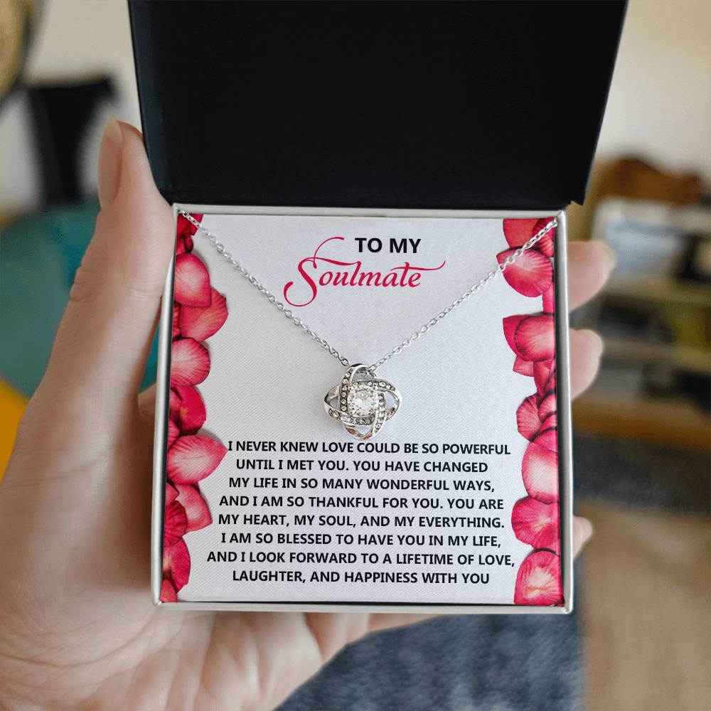 A hand holds an open box featuring the Soulmate-Until I - Love Knot Necklace, with its silver pendant gleaming elegantly. It is paired with a heartfelt message to a soulmate on a card decorated with red flowers.