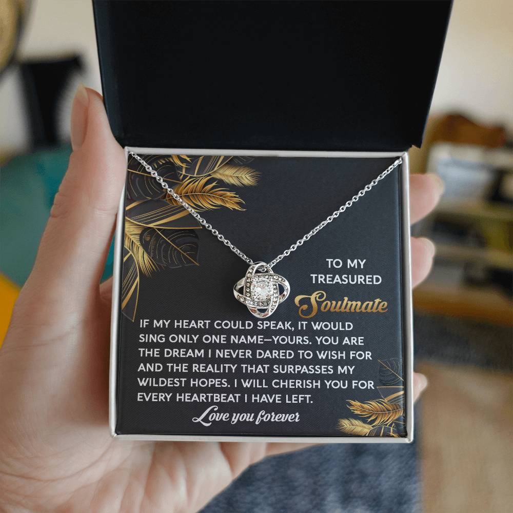 A hand holds the Soulmate-Wildest Hopes Love Knot Necklace adorned with cubic zirconia, displayed in a box with a heartfelt soulmate message. The pendant shines with a white gold finish.