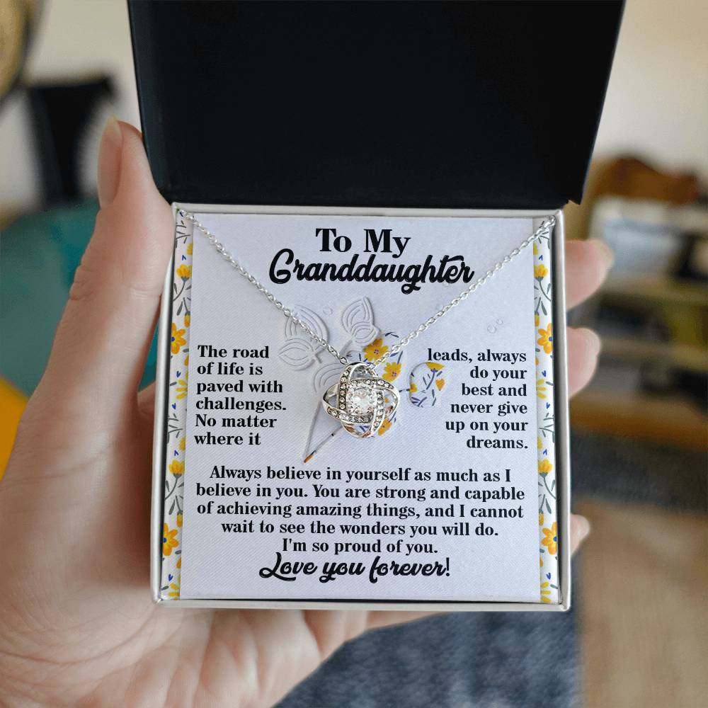 A hand holds an open gift box revealing a Granddaughter-On Your Dreams - Love Knot Necklace, adorned with sparkling cubic zirconia crystals and accompanied by a heartfelt message for a granddaughter.