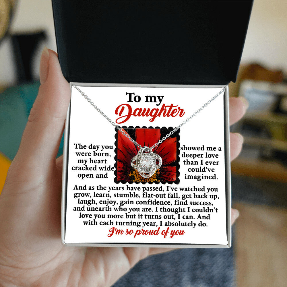 A hand holds an open jewelry box displaying the "Daughter-A Deeper Love - Love Knot Necklace" adorned with cubic zirconia crystals. Inside the cover is a heartfelt message addressed to "my daughter," expressing love, pride, and support, making it the perfect personalized gift.