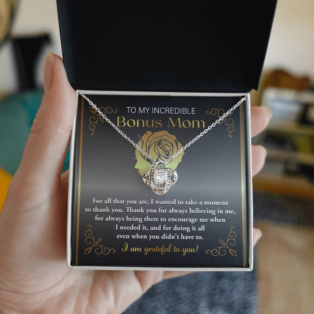Hand holding an open box containing a "To Bonus Mom, To Encourage Me - Love Knot Necklace" adorned with cubic zirconia crystals and accompanied by a message card that reads "To My Incredible Bonus Mom," expressing gratitude and appreciation.