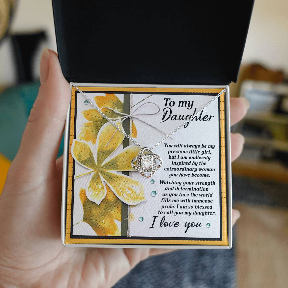 A person holds an open gift box containing the Daughter-Blessed To Call - Love Knot Necklace, adorned with cubic zirconia crystals. The box includes a heartfelt message to a daughter expressing love and admiration, with a floral design in the background. The necklace is available in either a white gold or yellow gold finish.